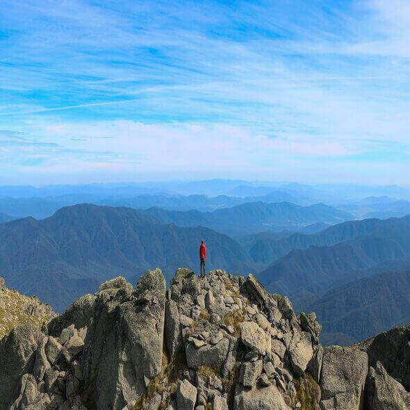 Person standing on rocky mountain peak overlooking vast Australian landscape under clear blue sky.