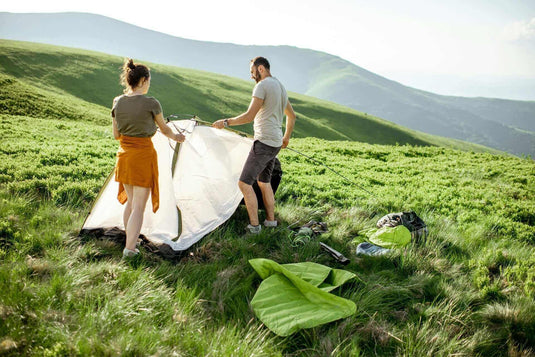 Couple setting up tent on a grassy hillside, planning a zero-waste camping trip. - Adventureco Blog