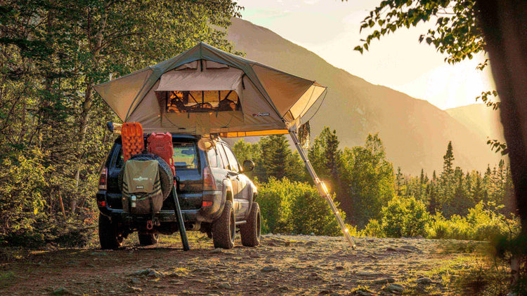 SUV with roof tent set up in a scenic forested camping spot with mountain view at sunset.