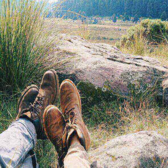 Hikers resting on a rock with their hiking boots on, enjoying a scenic view of nature with grassy hills and forest in the background