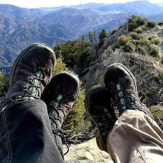 Hikers' shoes resting on a mountain peak with scenic view of green valley and hills, highlighting the need for sturdy hiking gear. - Adventureco Blog
