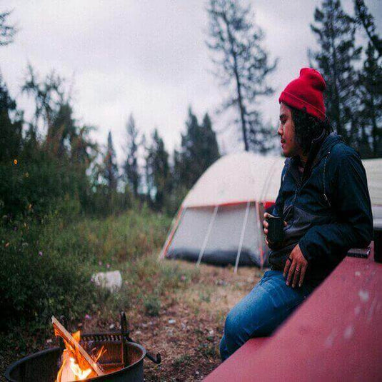 Camper enjoying a warm fire near a tent in a forest setting, wearing a red beanie and holding a mug.