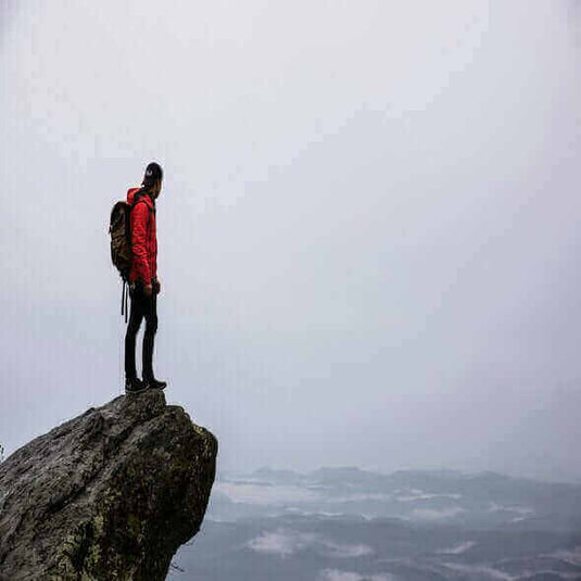 Hiker in red jacket standing on cliff edge overlooking misty landscape, illustrating responsible hiking and adventure in nature. - Adventureco Blog