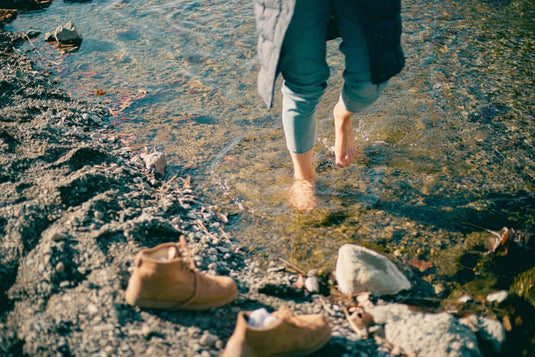 Person walking barefoot in water while barefoot shoes for flat feet are left on the shore. - Adventureco Blog