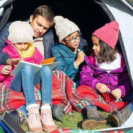 Family reading a book together while camping in a tent, kids wearing warm hats and father pointing at the book.