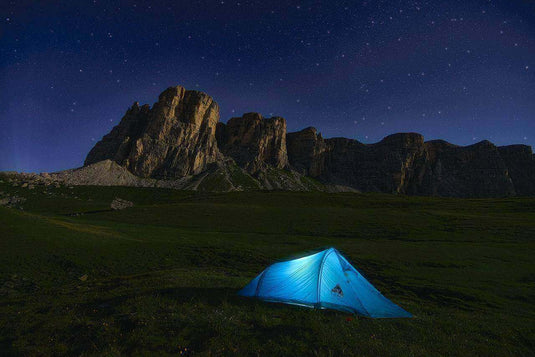 A blue tent illuminated under the starry night sky in a scenic mountain landscape, perfect for a comfy night of stargazing while camping.