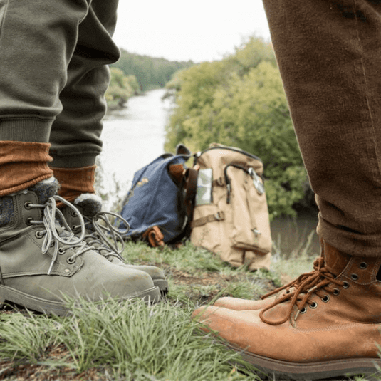 Two pairs of hiking boots on grass near a river, ideal for discussions on preventing blisters in barefoot shoes. - Adventureco Blog