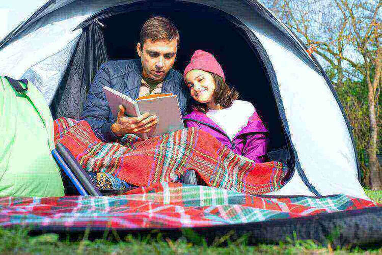 Parent and child reading a book inside a tent during camping trip for an environment-friendly adventure.