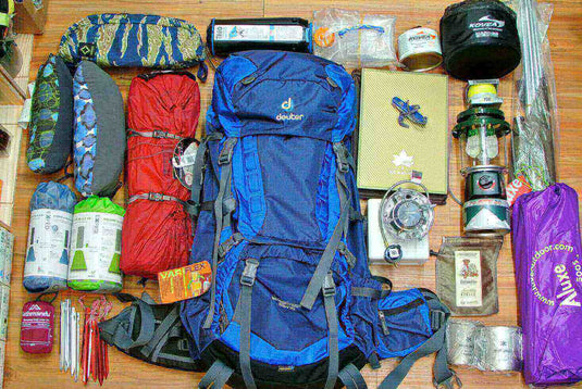 Camping gear including a blue backpack, sleeping bags, cooking equipment, and other essentials organized on a wooden floor.