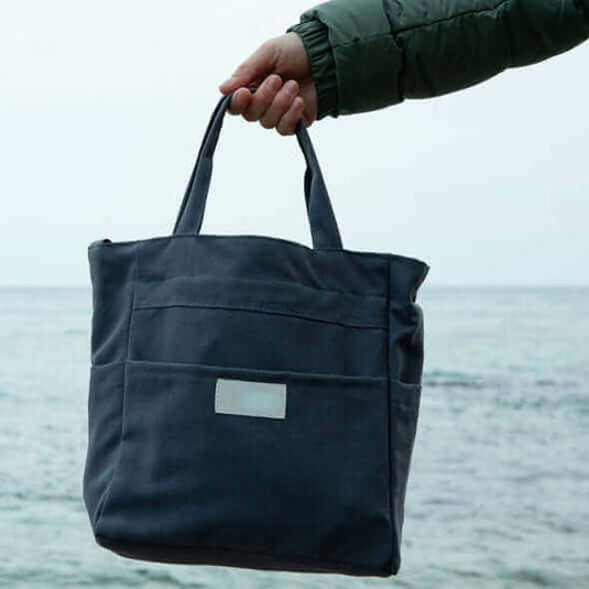 Hand holding a recycled sail bag against a scenic ocean background, promoting sustainable travel gear.