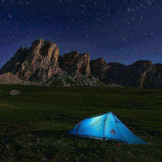 Blue tent set up at a campsite with mountainous background under a starry night sky, illustrating camping trip outdoors.