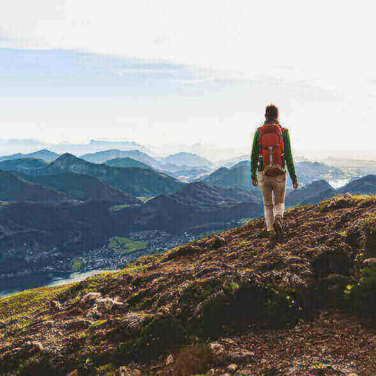 Hiker with a red backpack walking on a mountain trail, overlooking a scenic valley, emphasizing outdoor appreciation and low environmental impact. - Adventureco Blog