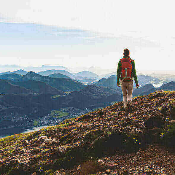 Hiker with a red backpack walking on a mountain trail, overlooking a scenic valley, emphasizing outdoor appreciation and low environmental impact.