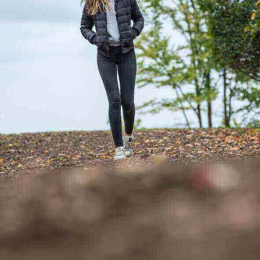 Woman wearing minimalist shoes and a jacket walking on a hiking trail surrounded by trees and nature