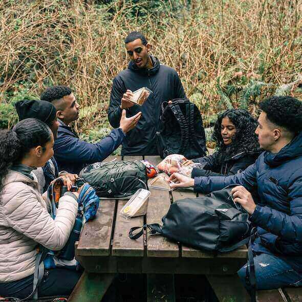 Family enjoying a meal at a picnic table during a day hike, surrounded by nature with essential hiking gear and food.