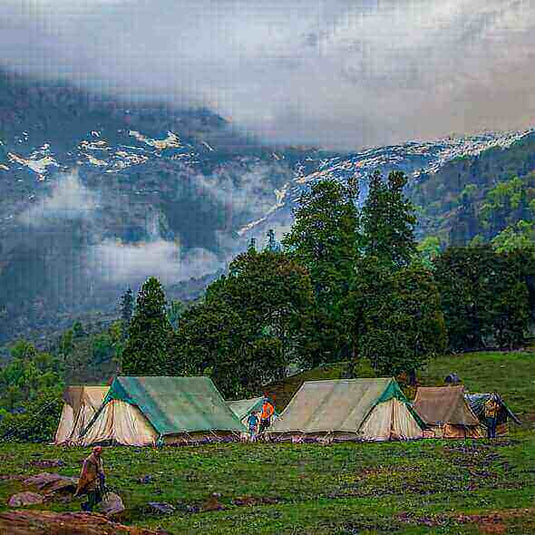 Campers setting up tents in a lush green meadow with misty mountains in the background on a rainy day.