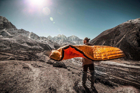 Camper holding an orange sleeping pad in a mountainous outdoor setting under the sun.