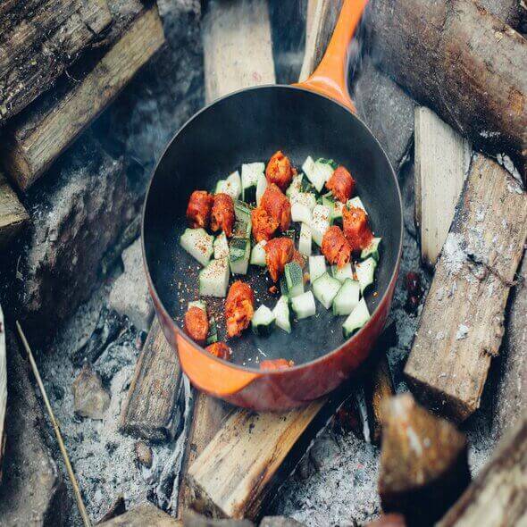 Cooking vegetables and meat in a skillet over a campfire with wooden logs for an outdoor meal.