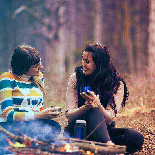 Two women enjoying coffee by a campfire in the forest while discussing clean energy for camping.