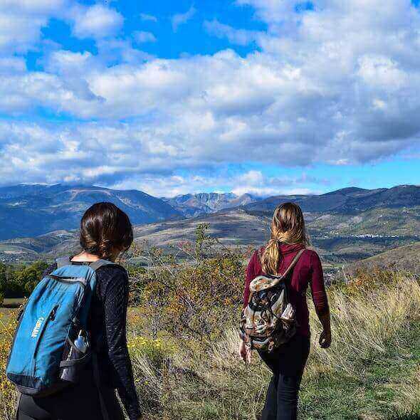 Two hikers with backpacks walking on a trail with scenic mountain views under a blue sky.