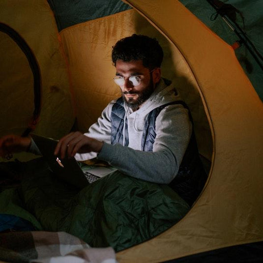 Camper in a tent using a laptop at night, preparing for rainy weather during camping trip