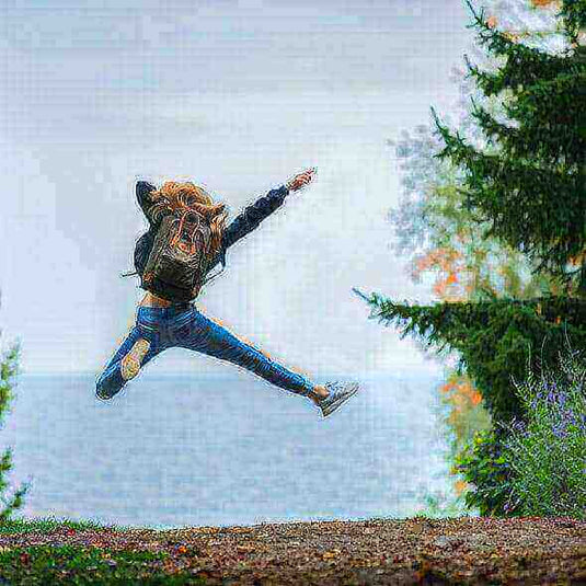 Woman jumping joyfully near a lake while backpacking, surrounded by lush greenery.