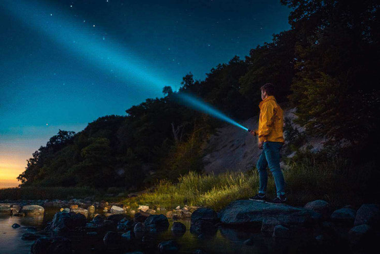 Camper illuminating the night with a powerful flashlight by the river, essential lighting tool for camping and outdoor trips