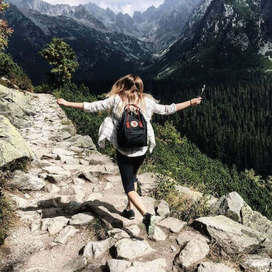 Woman hiking on a rocky path surrounded by mountains and forests, enjoying nature with a backpack.