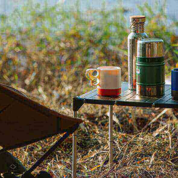 Travel mugs and tumblers on an outdoor table, showcasing zero waste insulated containers for hot and cold beverages during a camping trip.