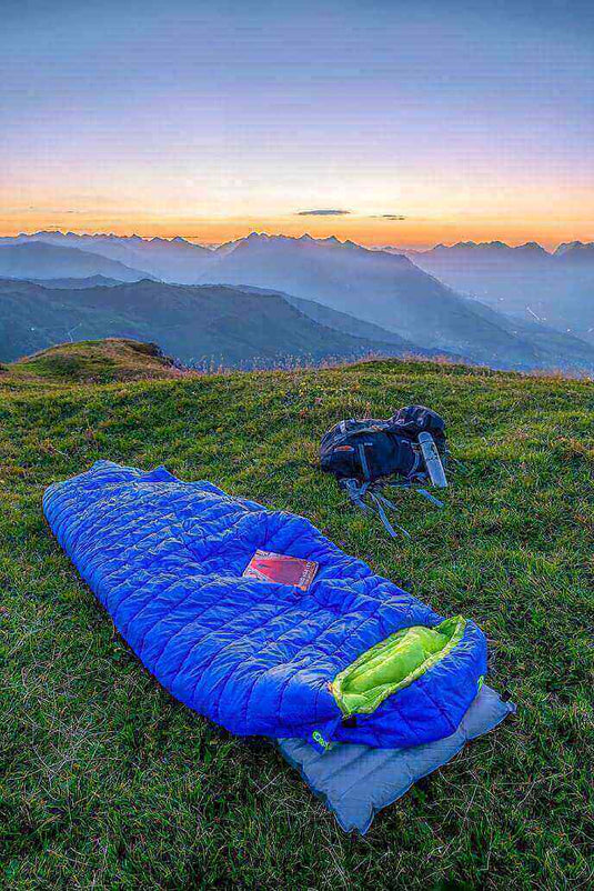 Blue sleeping bag on grassy hill at sunrise with hiking gear beside it and panoramic mountain view in the background.