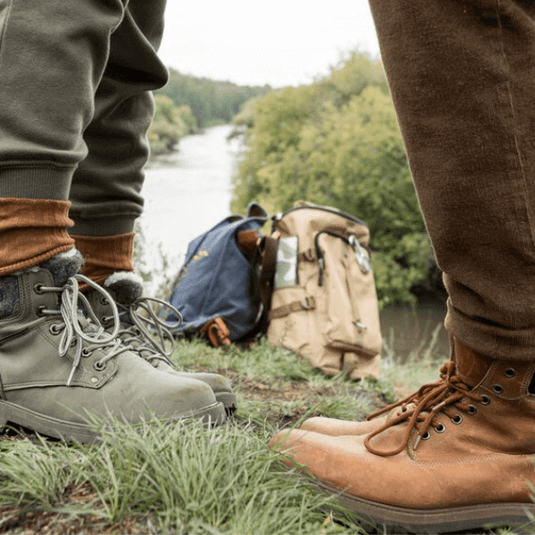 Two pairs of hiking boots on grass near a river, ideal for discussions on preventing blisters in barefoot shoes. - Adventureco Blog