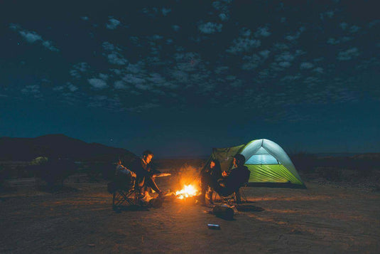 Campers sitting around a campfire near a glowing tent under a starry sky, enjoying a peaceful night in nature. - Adventureco Blog