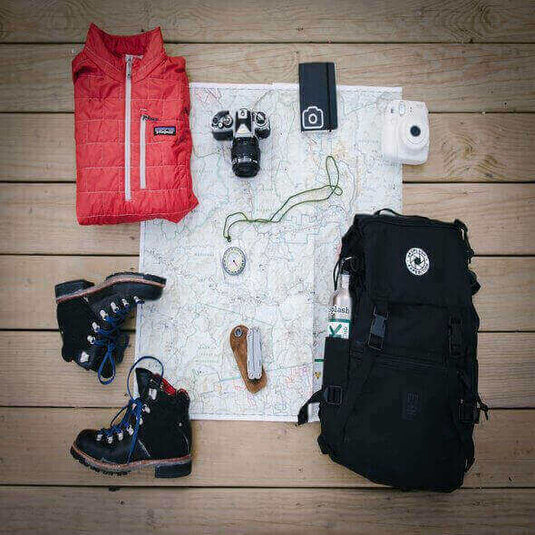 Sustainable hiking and camping gear including jacket, boots, backpack, map, camera, compass, and knife arranged on a wooden surface. - Adventureco Blog