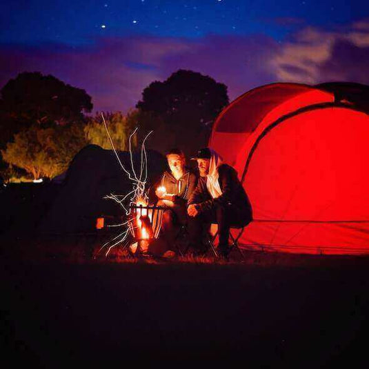 Campers sitting around a warm campfire at night, beside a brightly lit red tent under a clear, starry sky in the Australian outback.