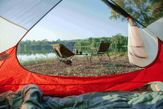 View from inside a red tent showing two camping chairs by a lakeside, surrounded by nature - minimalist camping setup.