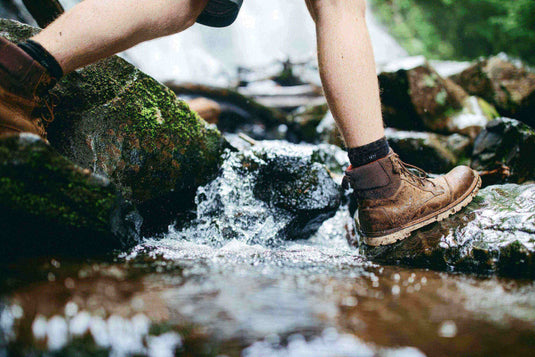 Person wearing sturdy hiking boots stepping on rocks across a stream in a forest.