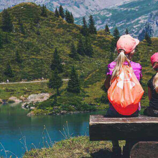 Children hiking in the mountains with backpacks, sitting on a bench overlooking a scenic lake and lush green landscape.