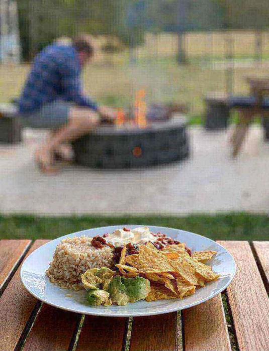 Plate of nachos with rice, avocado, and toppings on a wooden table with a person sitting by a fire pit in the background.