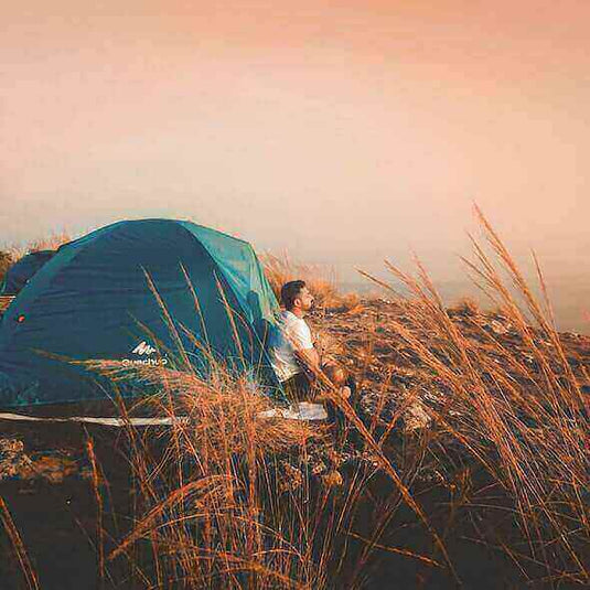 Beginner solo camper sitting beside tent on grassy hill at sunrise.