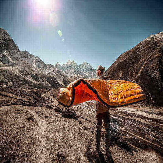 Person holding an orange sleeping bag in a mountainous outdoor setting.