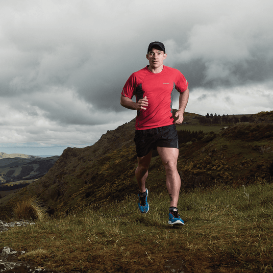 Man wearing red baselayer top and black shorts running on a trail in a mountainous area.