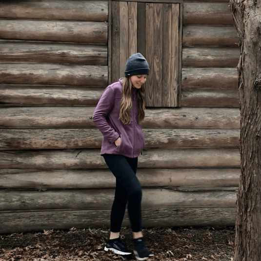 Woman in purple jacket and beanie walking outdoors in front of a rustic cabin.