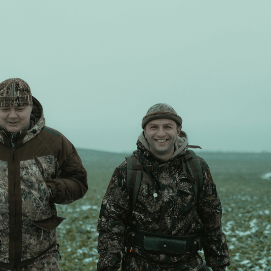 Two men wearing camouflage jackets and hats, standing outdoors in a field on a cold day.