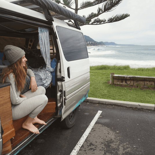 Woman in grey baselayer sitting in a van by the seaside enjoying the view.