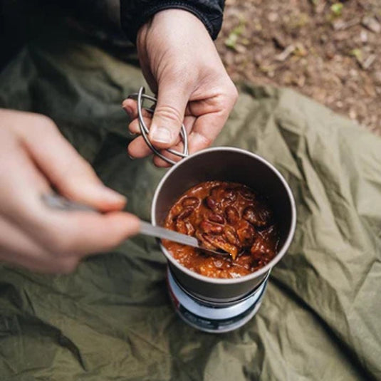 Person pouring Adventureco hiking meal into a cooking pot at a campsite with scenic background