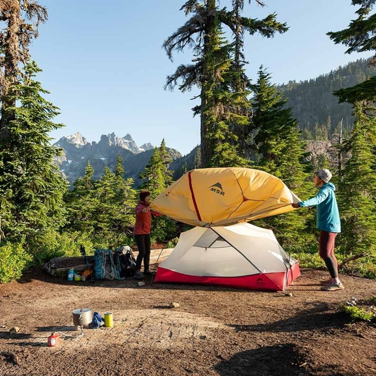 Campers setting up a durable AdventureCo tent in a scenic forest with mountains in the background.