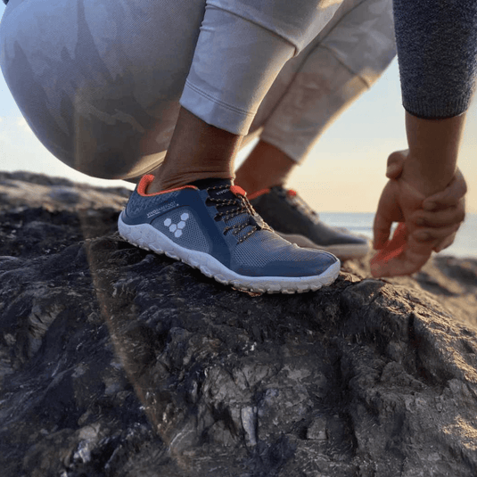 Person wearing blue Vivobarefoot shoes, crouching on rocky terrain at sunrise, showcasing lightweight and minimalist design for comfort and performance.