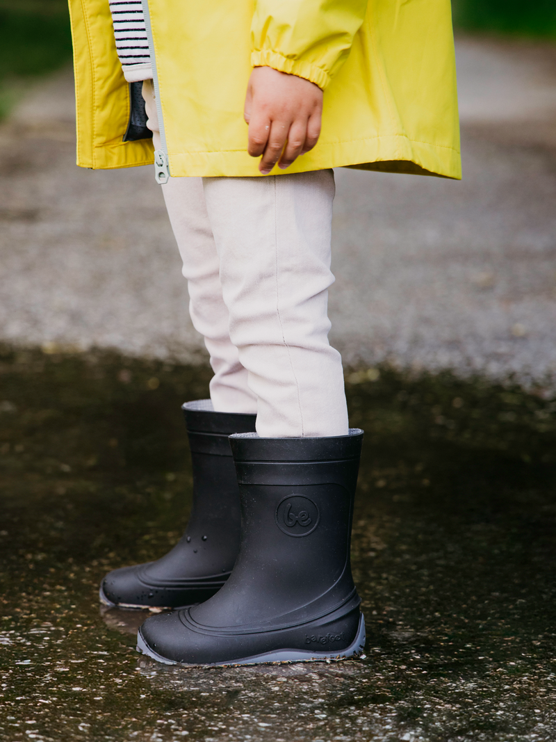 Load image into Gallery viewer, Child wearing Be Lenka Kids Barefoot Rain Boots in black, standing on a wet surface in a yellow rain jacket.
